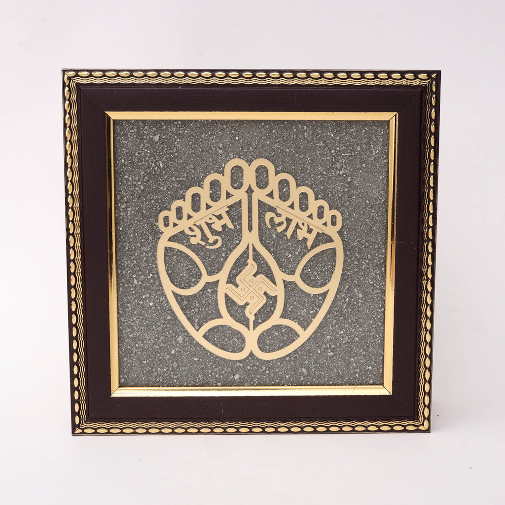 Gold laser-cut Charan Paduka sacred feet with Shubh Labh Devanagari inscription and Swastik symbol on charcoal stone texture background in dark wood decorative frame
